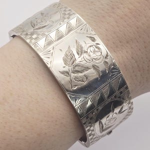 May include: A silver-colored hinged bangle bracelet with engraved floral and geometric patterns. The bracelet features a central floral design with leaves and a flower, surrounded by geometric shapes and decorative borders. The clasp is visible on the side.