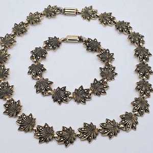 May include: A gold-coloured necklace and bracelet set. The jewellery features a repeating pattern of stylized leaf-shaped links. The necklace and bracelet are displayed on a white background, showcasing the intricate design and metallic finish.