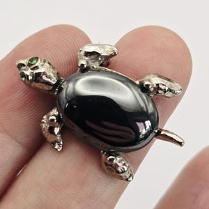 May include: A silver turtle brooch with a shiny black shell and green gemstone eyes. The turtle's head, feet, and tail are detailed with textured silver. A small, decorative accessory.