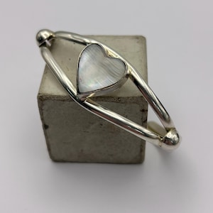 May include: A silver-toned bracelet with a heart-shaped mother-of-pearl inlay. The bracelet has a curved design with rounded end caps. The heart inlay is centered within the bracelet's frame, creating a delicate and elegant piece of jewelry.