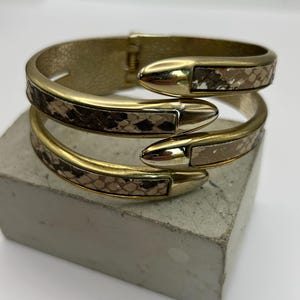 May include: A gold-coloured metal cuff bracelet with a snake-skin pattern. The bracelet has a hinged clasp and features a wrap-around design with pointed end caps. The snake-skin pattern is in shades of brown, tan, and black.