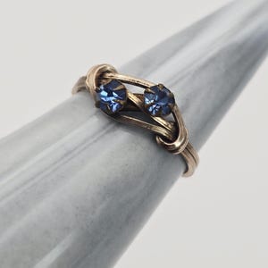 May include: A gold-toned ring featuring two round, blue gemstones. The ring's design incorporates a delicate, interwoven pattern, with the gemstones set prominently at the center. The ring is displayed on a light gray surface.