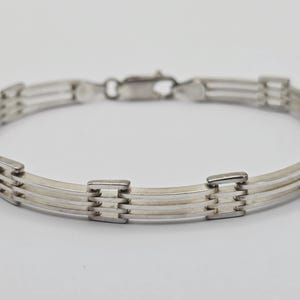 Puede incluir: Una pulsera de metal plateado que presenta tres barras horizontales paralelas conectadas por pequeños eslabones rectangulares a intervalos regulares. La pulsera incluye un cierre de mosquetón en la parte superior para un ajuste seguro.