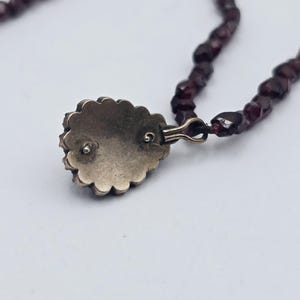 Puede incluir: Primer plano de un collar con un colgante con borde festoneado y una piedra preciosa de color rojo oscuro. El collar tiene un dise&ntilde;o de cuentas con peque&ntilde;as cuentas de color rojo oscuro. El colgante est&aacute; sujeto con un cierre dorado.