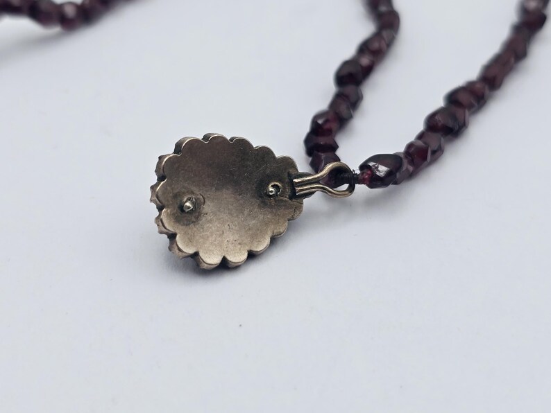 Puede incluir: Primer plano de un collar con un colgante con borde festoneado y una piedra preciosa de color rojo oscuro. El collar tiene un dise&ntilde;o de cuentas con peque&ntilde;as cuentas de color rojo oscuro. El colgante est&aacute; sujeto con un cierre dorado.