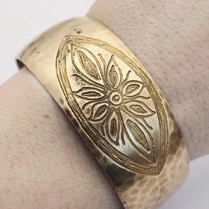 May include: A wide, gold-toned cuff bracelet with a hammered texture. The bracelet features an embossed floral design within an oval shape at the centre. The cuff is approximately 6.35 cm wide.