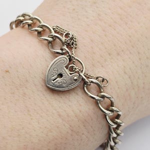 May include: Silver-toned charm bracelet featuring a heart-shaped padlock charm. The bracelet has a curb chain design with a small chain connecting the padlock to the main chain. The padlock has a keyhole design.