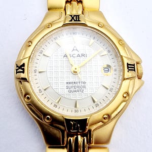 Puede incluir: Reloj de cuarzo Ascari dorado con esfera blanca y agujas doradas. La esfera del reloj presenta el nombre de la marca y las palabras "Ameretto Superior Quartz". Los números romanos marcan las posiciones de las 12, 3 y 9 en punto. La correa del reloj también es dorada.
