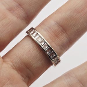 Op de afbeelding: Een zilveren ring met een rij vierkant geslepen, heldere edelstenen. De ring wordt op een vinger getoond, waarbij het ontwerp en de glans van de stenen worden benadrukt. De band is van gepolijst metaal.