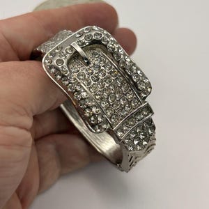 Può includere: Bracciale argentato con design a fibbia. La fibbia è tempestata di numerosi strass chiari e scintillanti. Il bracciale presenta un motivo inciso dettagliato sui lati. Un accessorio alla moda.