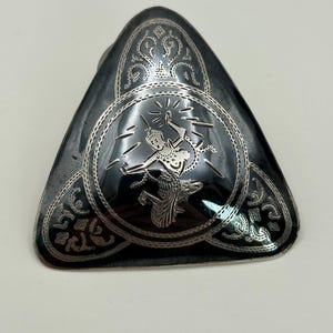May include: A triangular, silver-toned metal brooch with a black enamel background. The brooch features an engraved design of a dancing figure within a circular frame, surrounded by ornate patterns. The overall design is intricate and detailed.