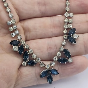 Puede incluir: Un collar plateado con piedras preciosas claras y azul oscuro. El collar presenta piedras claras de talla redonda y piedras azul oscuro de talla marquesa dispuestas en un diseño floral. El collar se sostiene en una mano.