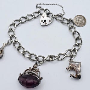 May include: Silver charm bracelet with a chain link design. The bracelet features several charms, including a heart-shaped padlock, a coin, a claw holding a pearl, a faceted purple gem, and a small boot.