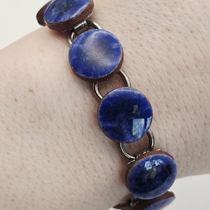 Puede incluir: Una pulsera con piedras redondas de color azul oscuro con apariencia veteada, engastadas en marcos marrones y conectadas por eslabones plateados. La pulsera se muestra en una muñeca, mostrando su diseño y color.