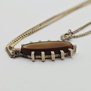 May include: A gold-toned necklace with a brown oval pendant. The pendant has a rectangular gold frame with vertical bars. The chain is a fine link style, and the pendant is centered on the chain.