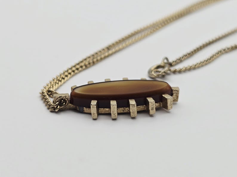 May include: A gold-toned necklace with a brown oval pendant. The pendant has a rectangular gold frame with vertical bars. The chain is a fine link style, and the pendant is centered on the chain.