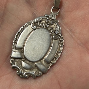 May include: A silver pendant with an oval centre and decorative scrollwork. The pendant has a bail for a chain and a detailed border. The design is vintage and elegant, with a classic style.