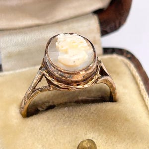 May include: A vintage gold ring with an oval cameo. The cameo features a carved profile of a woman in cream and is set in a gold bezel. The ring is displayed in a velvet-lined jewellery box.