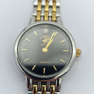May include: A silver and gold-tone Beverly Hills Polo Club quartz watch. The watch has a black face with gold-tone hands and markers. The watch band is a combination of silver and gold-tone links.