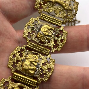 May include: A vintage gold-toned bracelet featuring ornate filigree links. Each link showcases a detailed design, including a sword and a lion's head. The bracelet's intricate patterns and aged appearance suggest it is an antique or a piece of costume jewelry.
