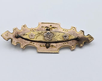 May include: An antique brooch with a floral design. The brooch features a central gold-toned oval with a flower and leaf motif, framed by a decorative, gold-toned and rose-toned metal border. The brooch is approximately 3 inches long.