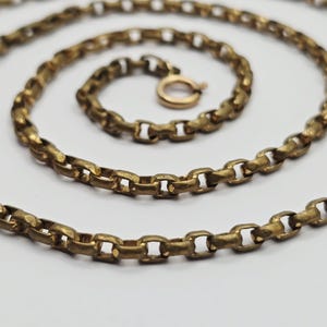 Puede incluir: Un collar de cadena de metal en tono latón que presenta una serie de eslabones redondos interconectados. La cadena está dispuesta en un patrón en espiral e incluye un cierre de resorte circular en el extremo. La superficie metálica tiene un acabado mate.