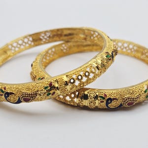 May include: Two gold bangles with detailed patterns. The bangles have a textured surface and are decorated with colourful accents, including red, green, and blue details. The design includes peacock motifs and openwork patterns.