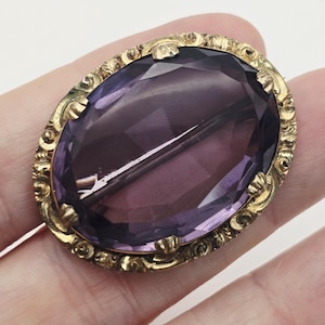 Puede incluir: Broche ovalado antiguo con una gran gema púrpura facetada engastada en un marco dorado y ornamentado. El marco tiene un diseño detallado en relieve. El broche tiene un cierre de alfiler en la parte posterior.