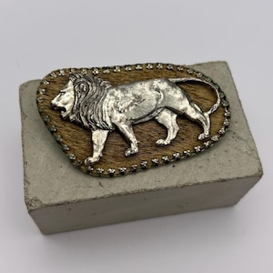 May include: A silver-colored lion belt buckle. The lion is depicted in a walking pose on a brown background, surrounded by a decorative border. The buckle is rectangular and appears to be made of metal.