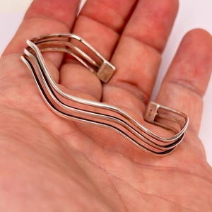 May include: A silver-toned cuff bracelet with a wavy design. The bracelet features four parallel bands that curve and undulate across the wrist. The open-ended design allows for easy wear. The bracelet is shown on a light skin tone.