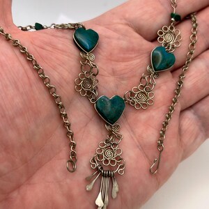 May include: A silver-toned necklace featuring heart-shaped turquoise stones. The necklace has a delicate chain and intricate wirework with floral and spiral designs. The pendant has a cluster of dangling silver elements.