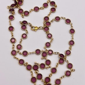 Puede incluir: Un collar de cadena en tono dorado que presenta una serie de piedras de vidrio magenta circulares engastadas. El collar está construido con eslabones metálicos individuales que conectan cada engaste y un cierre de mosquetón.