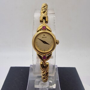 Puede incluir: Un reloj de pulsera Seiko dorado con esfera crema y agujas negras. El reloj tiene una esfera redonda con una pequeña corona y está adornado con dos piedras preciosas rojas. La correa del reloj tiene un diseño trenzado.