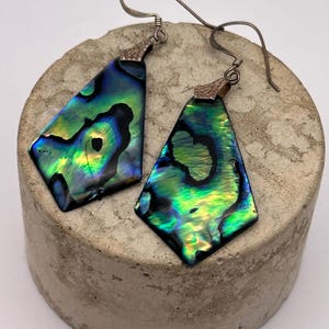 May include: A pair of iridescent abalone shell earrings. The earrings are diamond-shaped with a silver hook. The abalone shell displays a vibrant mix of green, blue, and black colors. The earrings are displayed on a gray, textured surface.