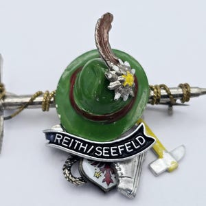 Pode incluir: Broche detalhado com um chapéu verde com pena, uma faixa com a inscrição "REITH/SEEFEELD" e um pequeno martelo. O broche tem uma base de metal prateado com detalhes dourados e um emblema de escudo.