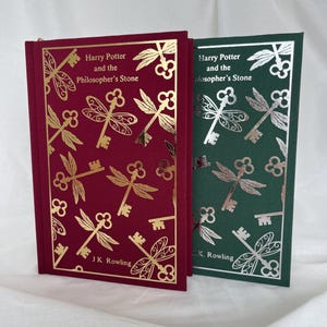 May include: Two hardcover books, "Harry Potter and the Philosopher's Stone," in burgundy and forest green. The burgundy book has gold foil dragonfly and key designs, while the green book has silver foil. Both books feature the author's name, J.K. Rowling.