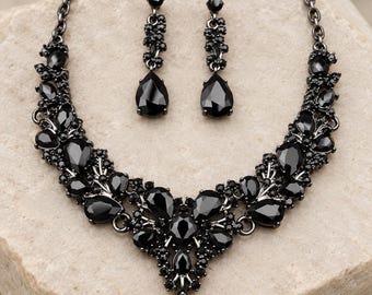May include: A jewelry set consisting of a necklace and a pair of drop earrings, both crafted from dark gunmetal-toned metal and adorned with numerous faceted black crystals in various shapes, including prominent teardrop-cut stones.