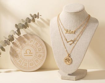 May include: A gold-toned layered necklace set displayed on a beige mannequin bust. The top layer features the word Libra in an old English font, the middle layer shows a constellation charm, and the bottom layer displays a circular scale pendant.