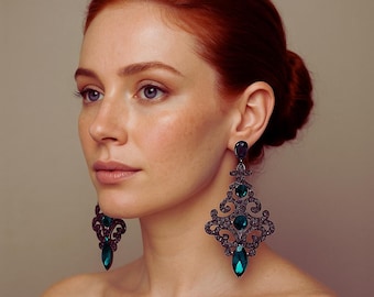 May include: A woman with red hair and blue eyes wears large, ornate chandelier earrings. The earrings are dark gray with teal-colored teardrop and round gemstones. The earrings have a filigree design.