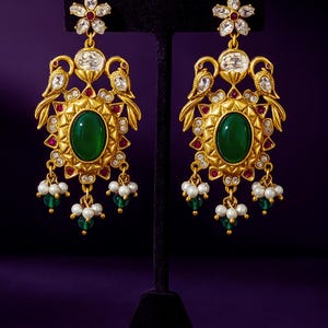 May include: Gold-toned earrings with an oval green gemstone center, surrounded by red and clear stones. The earrings feature bird and flower designs, with dangling pearls and green beads. The earrings are displayed on a black stand against a purple background.