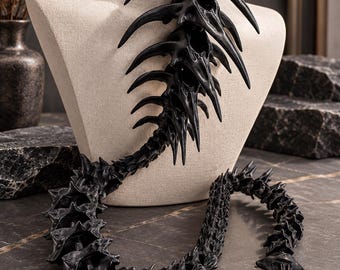 May include: A black leather choker featuring a long, articulated spine-like pendant made of dark, textured bone-shaped segments that drape downward, ending in a pointed, detailed tip with subtle metallic highlights.