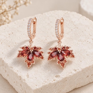 May include: A pair of rose gold hoop earrings featuring a dangling maple leaf charm. Each leaf is adorned with deep red pear-shaped stones and smaller orange accent stones. The hoop portion of each earring is encrusted with tiny clear white stones.