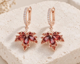 May include: A pair of rose gold hoop earrings featuring a dangling maple leaf charm. Each leaf is adorned with deep red pear-shaped stones and smaller orange accent stones. The hoop portion of each earring is encrusted with tiny clear white stones.