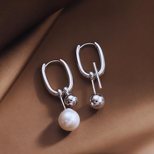 May include: Two silver hoop earrings with a modern design. One earring has a white pearl pendant, the other a silver ball. The earrings have a polished finish. The background is a warm, neutral tone, creating a contrast with the silver.