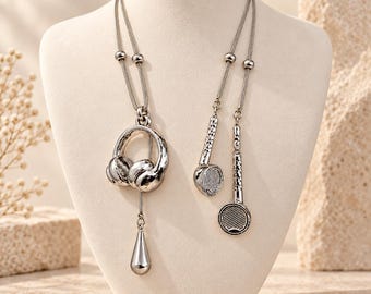 May include: A silver-toned necklace featuring a pendant shaped like a pair of headphones with a dangling teardrop charm, accompanied by two smaller dangling charms shaped like circular audio speakers, all suspended from a thin metallic chain.