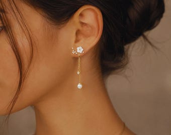 May include: A gold-toned earring featuring a white flower design with dangling chains and a pearl. The earring is adorned with small crystals and is worn on the ear. The jewelry is delicate and elegant.