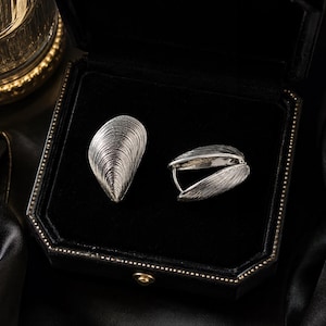 May include: A pair of silver earrings shaped like mussel shells, featuring detailed radial ridges and a textured metallic finish. One earring is displayed flat, while the other is shown from the side, revealing a hinged hoop mechanism.
