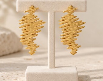 May include: A pair of gold-toned earrings featuring an elongated, jagged, zigzag silhouette. The surface of each earring is textured with horizontal, raised lines and small, evenly spaced dots, creating a dynamic, sharp-edged, abstract shape.
