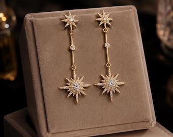 May include: A pair of gold-toned drop earrings featuring a small star-shaped stud at the top, connected by a thin vertical bar to a larger, multi-pointed star pendant. Both star elements are encrusted with small, clear, sparkling stones.