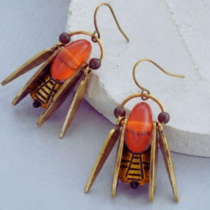 May include: A pair of insect-themed earrings. Each earring features an orange-red oval bead at the centre, framed by brass-toned metal wings and a detailed abdomen. The earrings have a hook closure.